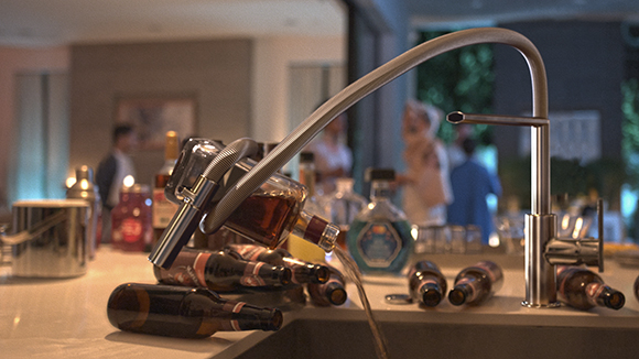 A glass bottle pouring liquid into a sink next to a silver faucet and various liquor bottles.