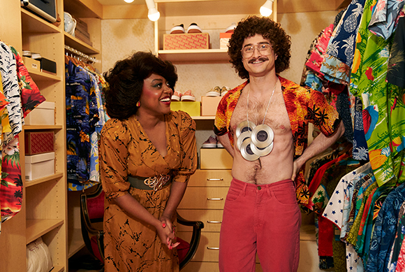 A smiling woman in an orange dress looks at a man wearing an open tropical shirt and a record necklace.