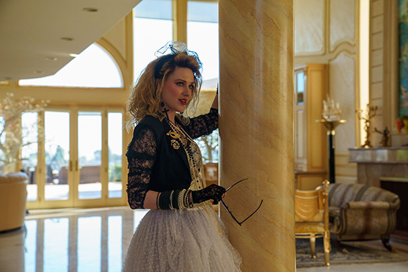 A woman with blonde hair wearing a black lace top and white tulle skirt poses behind a large marble column.