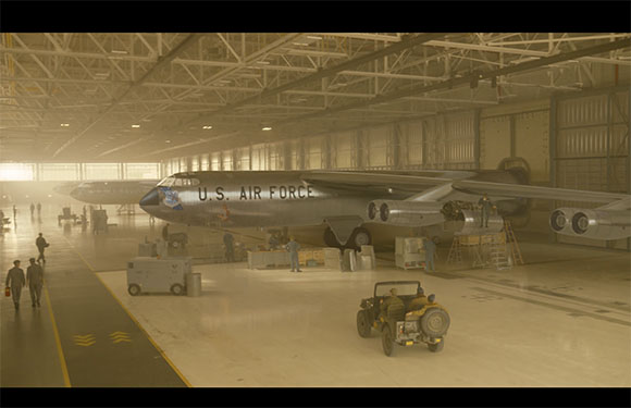 A large U.S. Air Force jet parked inside a vast, brightly lit aircraft hangar with personnel and vehicles.