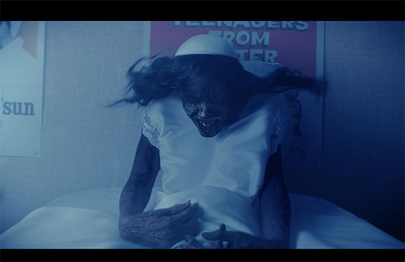 A person with dark, decaying skin and long hair wearing a white dress sits on a bed in a blue-tinted room.