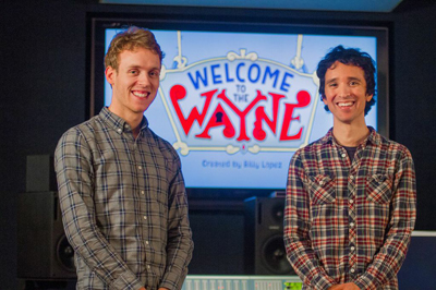 Two men in plaid shirts smiling in front of a screen showing the Welcome to the Wayne logo.