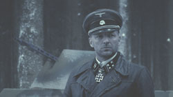 A man in a dark military uniform and peaked cap stands before a blurred background.