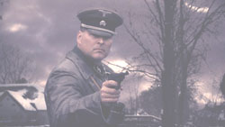A man in a dark military uniform and peaked cap points a handgun toward the camera.