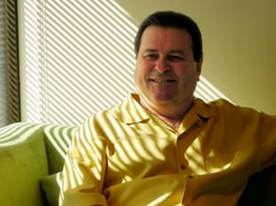 A smiling man wearing a yellow button-down shirt sits on a green sofa with sunlight through blinds.