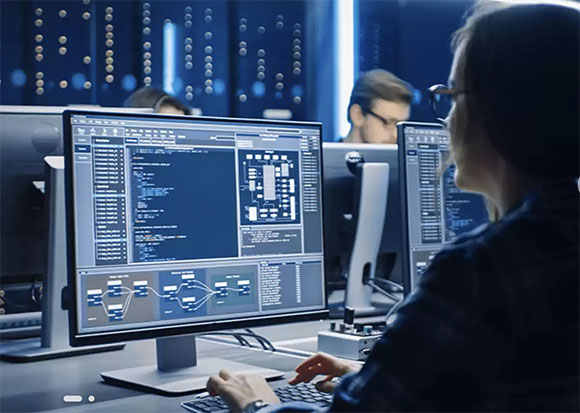 A person sits at a desk viewing computer monitors displaying lines of code in a dimly lit room.