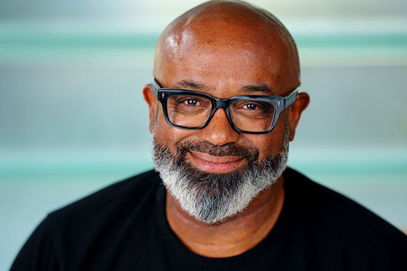 A smiling Black man with a bald head, black-rimmed glasses, and a salt-and-pepper beard.