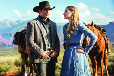A man in a grey coat and a woman in a blue dress stand before two horses in a desert landscape.