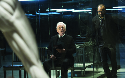 An older man sits in a chair while a younger man stands behind a glass partition in a dark room.