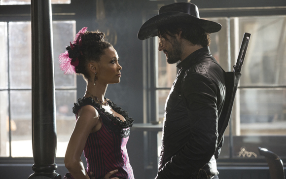 A woman in a pink striped corset and a man in a black leather jacket and cowboy hat face each other.