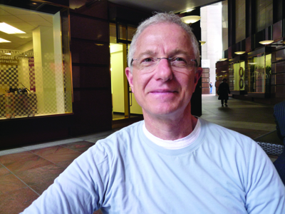 A man with short grey hair and glasses wearing a light blue t-shirt, smiling outdoors.
