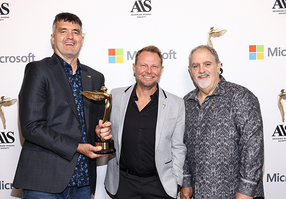 Three smiling men standing together, one holding a gold winged trophy against a white branded backdrop.