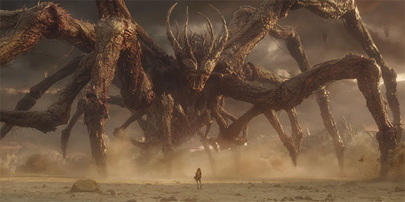 A massive, spindly-legged monster towers over a lone person in a vast, dusty desert.