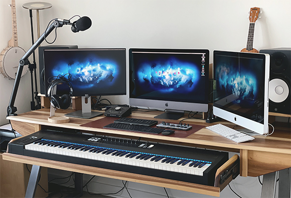 Music studio desk with three computer monitors, studio monitors, headphones, and a MIDI keyboard.