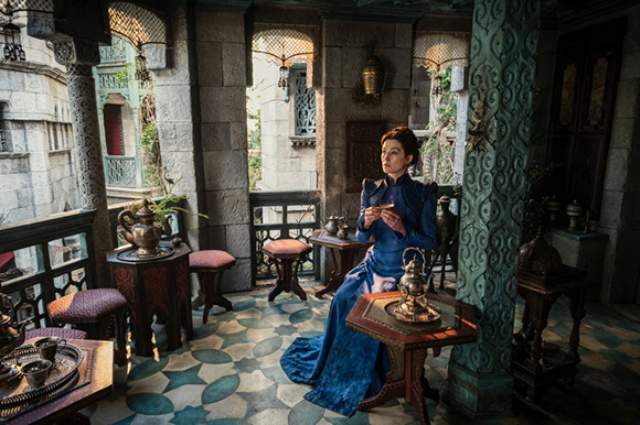 A woman in a long blue dress sits at a small table in a stone courtyard with patterned tile floors.