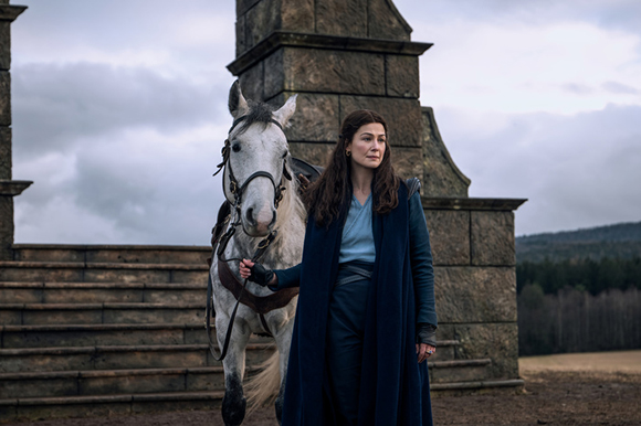 A woman with long dark hair in a blue coat stands beside a white horse in front of stone structures.
