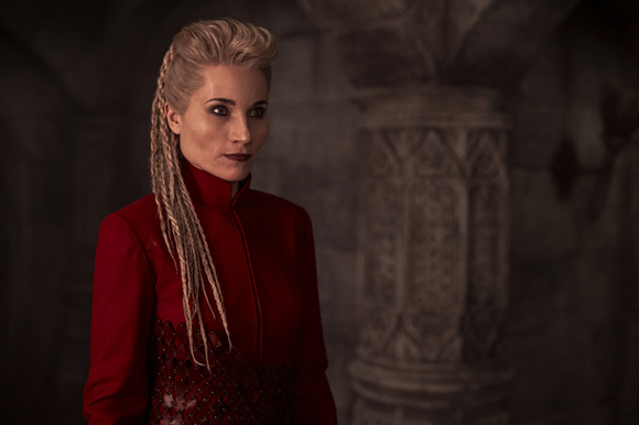 A woman with blonde dreadlocks and dark makeup wearing a red high-collared outfit against a dark stone background.