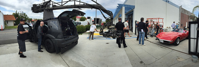 Film crew members working with a large camera crane and equipment near a black SUV and a red sports car.