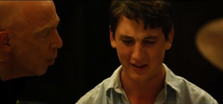 A bald man looks toward a younger man with a distressed facial expression in low lighting.