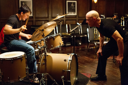 A drummer seated at a kit plays while a bald man in a black shirt leans toward him in a wood-paneled room.