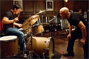 A man plays a drum kit while another man leans forward to watch him closely in a wood-paneled room.
