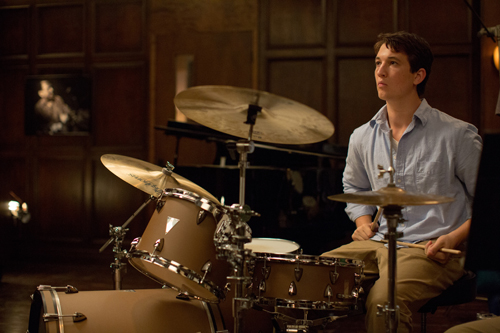 A young man in a light blue shirt sits behind a drum set in a dark, wood-paneled room.