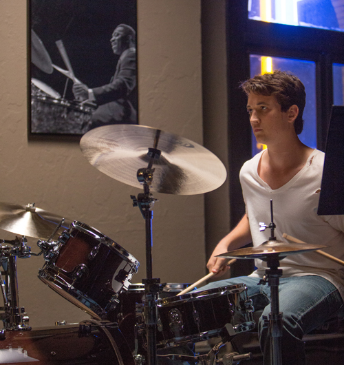 A man in a white shirt sits behind a drum kit, holding drumsticks and looking to the side.