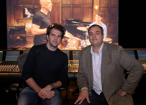 Two men sit in front of a large screen displaying a person at a piano in a studio.