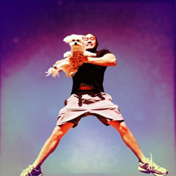 A man in a black shirt and shorts holds a small white dog while standing with legs spread wide against a purple sky.