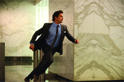 A man in a dark suit and blue shirt running through a building with marble walls.