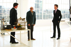 Three men in business attire stand in a bright white room near a table with a birdcage.