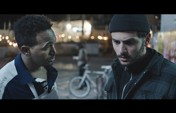 Two men stand close together in a dark outdoor area with blurred lights and a bicycle in the background.