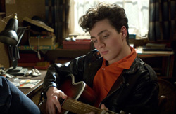 A young man with curly hair wearing a black leather jacket and orange shirt plays an acoustic guitar.