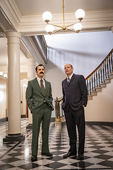 Two men in formal suits stand on a checkered floor in a grand hallway with a staircase.