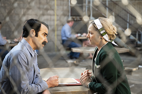 A man in a blue shirt and a woman in a green uniform sit at a table, viewed through a wire mesh.