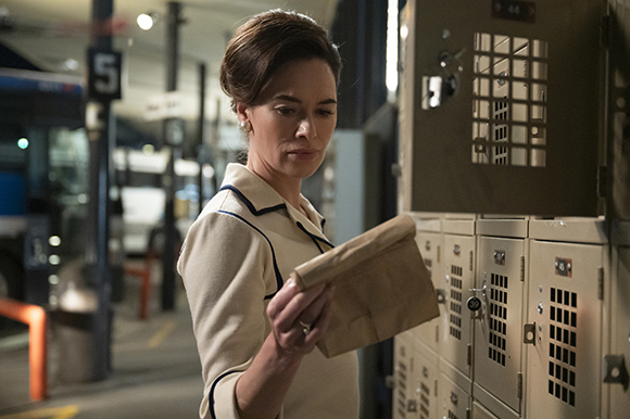 A woman in a white collared shirt examines a brown paper envelope next to a row of metal lockers.