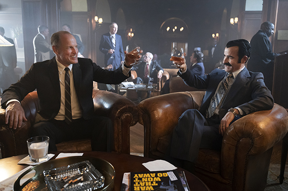 Two men in formal suits sit in leather armchairs, raising glasses of amber liquid in a toast.