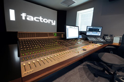 A large audio mixing console sits in a studio before computer monitors and a screen displaying the word factory.
