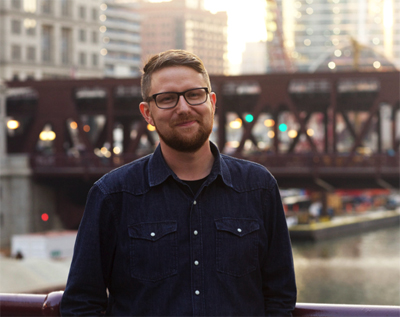 A man with short brown hair, glasses, and a beard wearing a dark denim shirt, smiling in an urban setting.