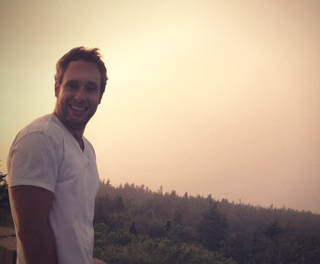 A smiling man in a white v-neck t-shirt standing against a hazy, sunset sky over a forest.