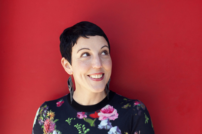 A smiling woman with short black hair and large earrings wearing a floral shirt against a red wall.