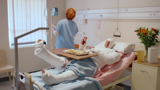 A patient in a hospital bed with legs and arms in white casts, while a nurse in blue scrubs stands nearby.