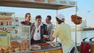 Three men talk to a vendor at an outdoor food stall by the water.