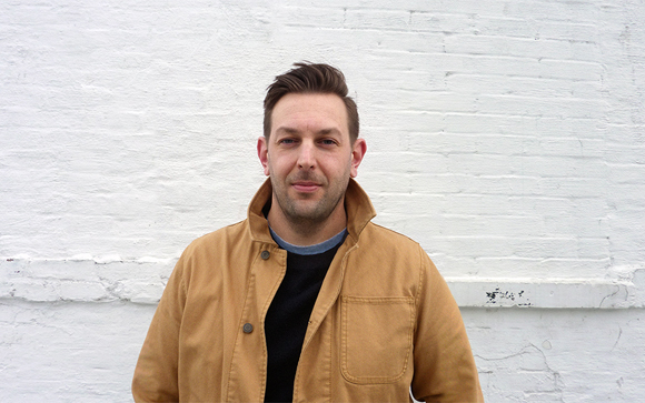A man with short brown hair wearing a tan jacket and black shirt against a white brick wall.