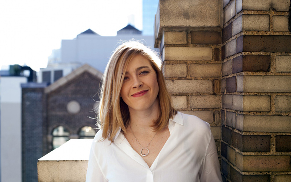 A woman with blonde hair wearing a white button-down shirt, smiling against a brick wall and city background.