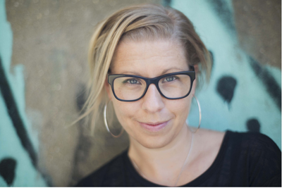 A blonde woman wearing black-rimmed glasses and large hoop earrings smiles against a blurred background.