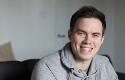 A smiling man with short dark hair wearing a grey hooded sweatshirt.