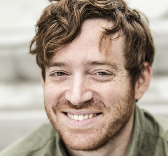 A close-up portrait of a man with wavy reddish-brown hair and a light beard, smiling at the camera.