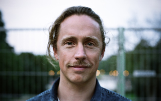 A man with light brown hair and a mustache smiling slightly against a blurred outdoor background.