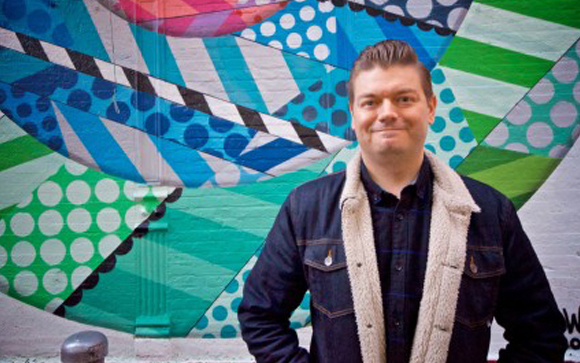 A man in a denim jacket with a shearling collar stands before a vibrant, geometric blue and green mural.